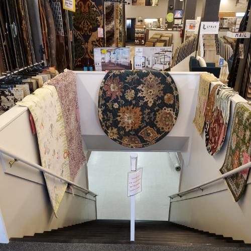 Flooring store carpet display with folded area rugs on shelves, a large round patterned rug on a stand, and multiple hanging pattern rugs; shelves and banners fill the background.